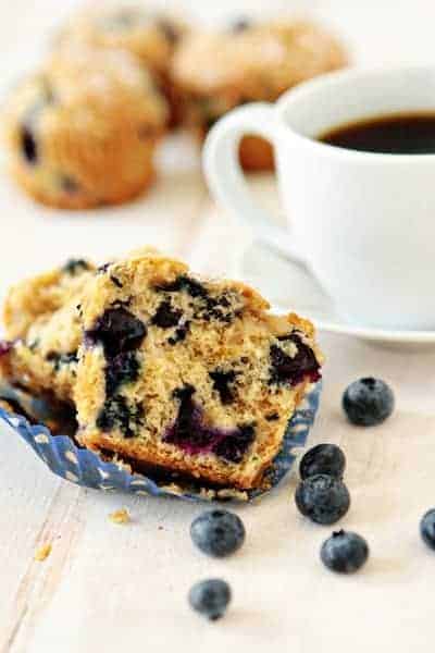 Blueberry lime muffin cut in half on a white surface in front of a cup of coffee and fresh blueberries