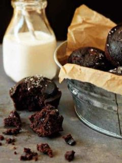 A galvanized bucket full of chocolate muffins next to a muffin broken in half with a glass of milk