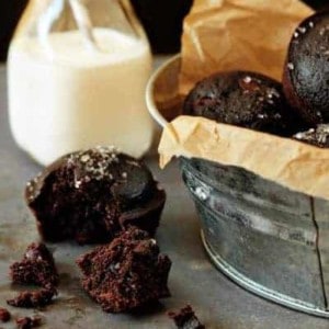 A galvanized bucket full of chocolate muffins next to a muffin broken in half with a glass of milk