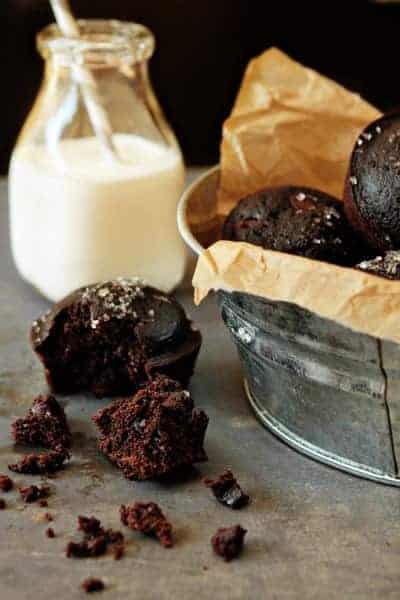 A galvanized bucket full of chocolate muffins next to a muffin broken in half with a glass of milk