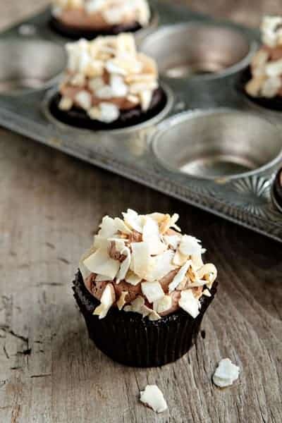A coconut mocha cupcake in front of a pan of cupcakes on a wood surface