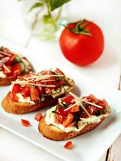 Three pieces of garlic tomato bruschetta on a white plate in front of a tomato
